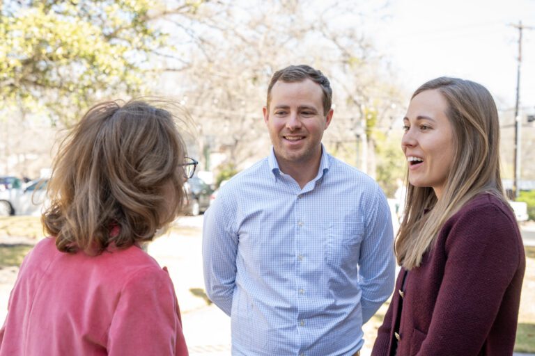 A woman wearing a pink jacket speaks with two newcomers, a man and a woman, outside on a sunny day near the church grounds.