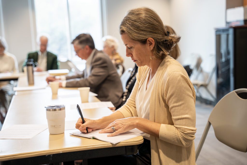 A woman takes notes during a Sunday Bible study session at a table, with other participants blurred in the background, reading and writing.
