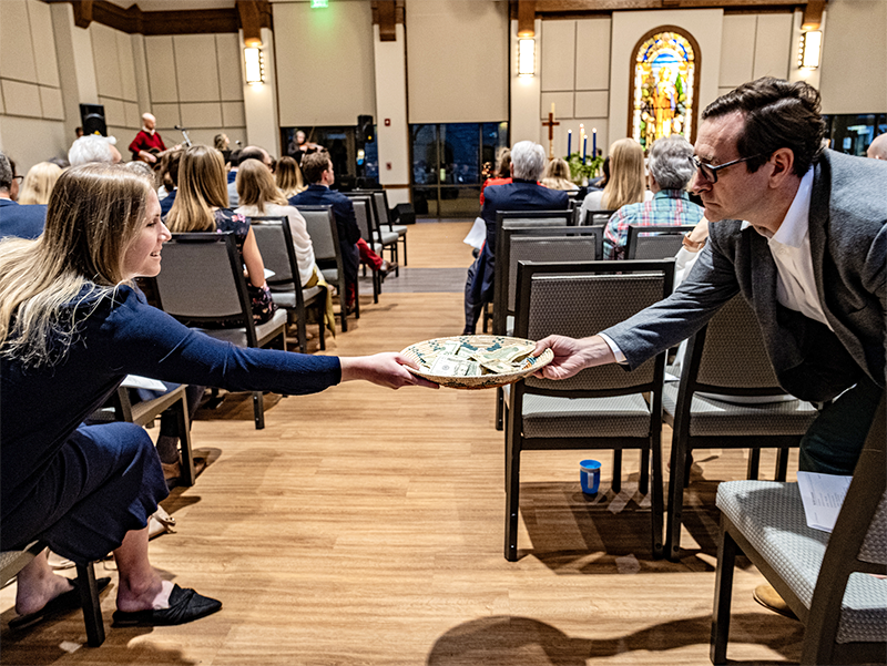 A woman reaches forward to place her donation into an offering plate filled with bills, handed to her by an usher during a church service.