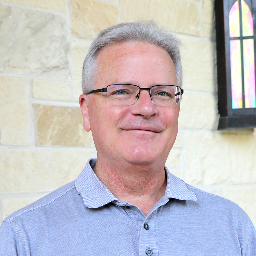 Portrait of a smiling man with short gray hair and glasses, wearing a light gray collared shirt, standing in front of a cream-colored stone wall.