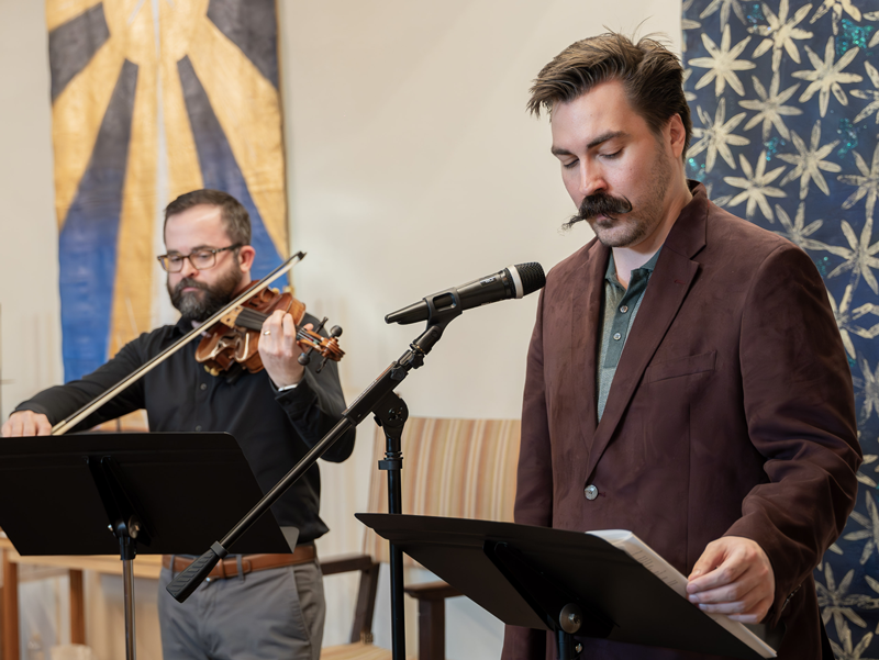 Two men performing during a church service—one playing the violin, the other reading into a microphone—at Good Shepherd on the Hill, in front of liturgical banners.