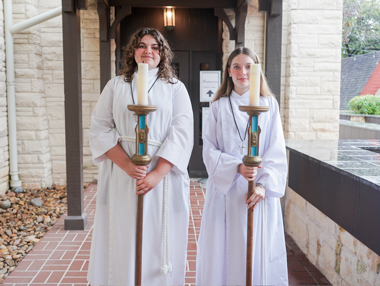 Two youth acolytes in white robes hold processional candles outside Good Shepherd Episcopal Church in Austin, Texas.